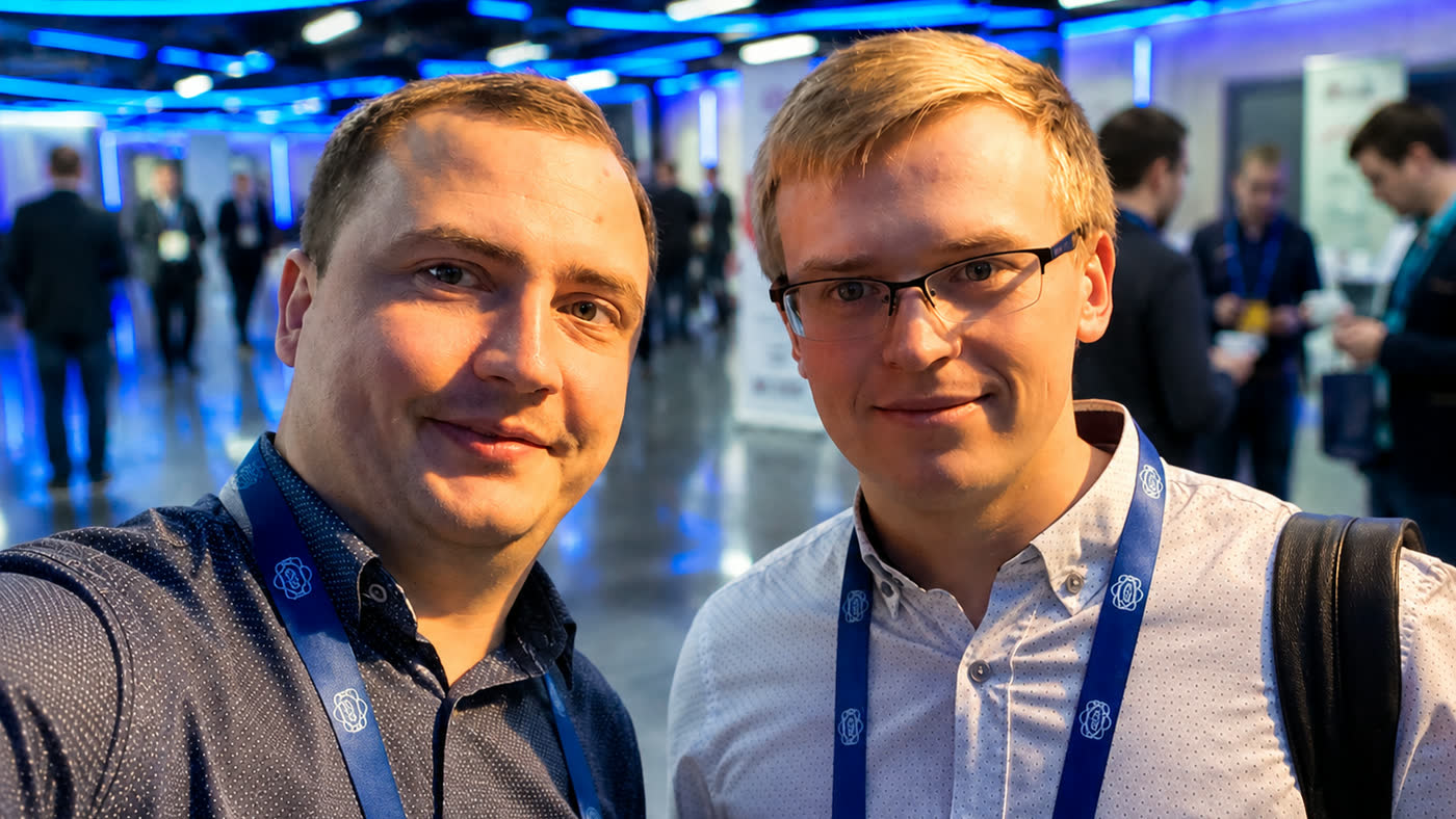 Two of the Fluvius team at a developer conference, lanyards on, the hall lit behind them