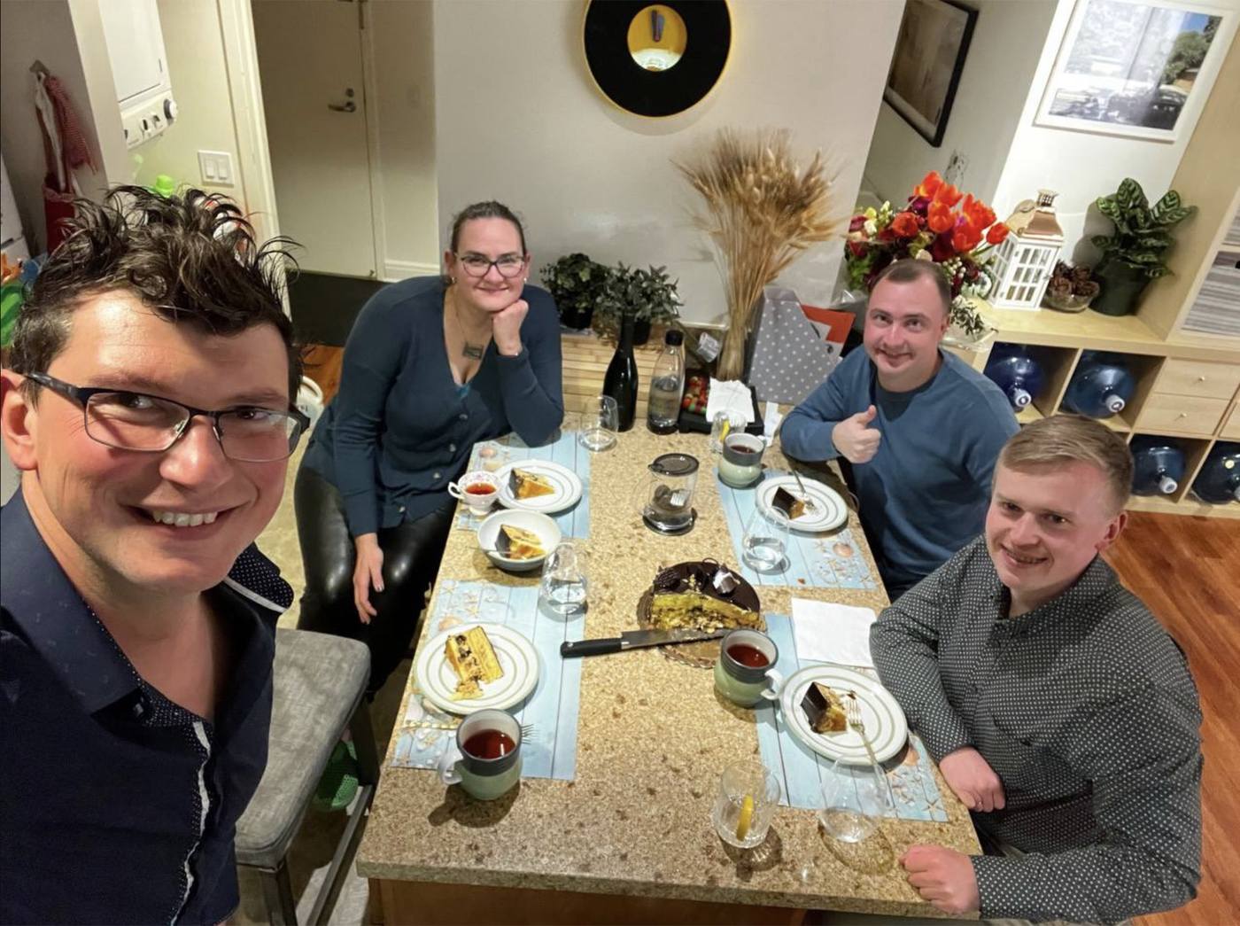 Four people around a kitchen table at the end of a working day, cake and tea in front of them, all turned toward the camera