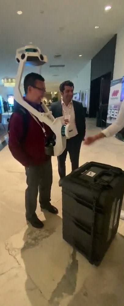 A spatial-scanning rig being worn on a show floor: a harness carrying a sensor head above the operator's shoulders and a battery pack across his chest, with a colleague watching beside him